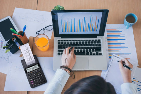 Person using laptop with graphs beside a Compact Desktop Printing Calculator With 12 Digit Display And Built In Printer For Tax And Currency Functions.