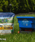 Handheld Lawn Spreader For Seed And Fertilizer, Easy Crank Broadcast Spreader in blue next to Black Beauty Ultra seed bag on grass