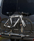 Bicycle mounted on a vehicle rack during rain, secured with Two Pack Retractable Ratchet Straps 2 Inch By 10 Foot Tie Down Straps