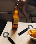 Ohiyoo metal bottle opener kit with black handle; two openers on a wooden table, one used to open a beer bottle.