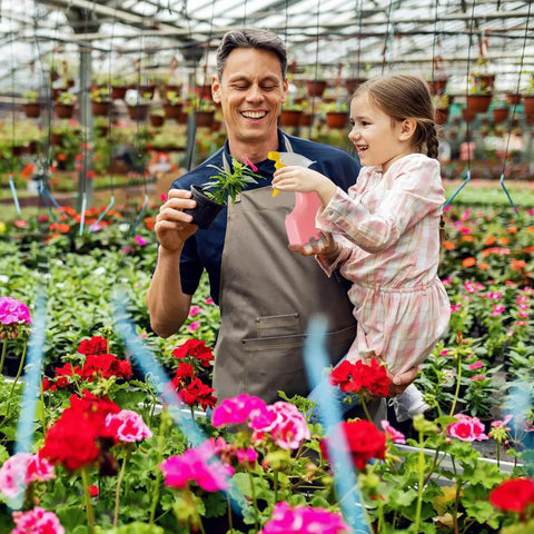 Adult and child in garden holding plant and watering can amidst flowers, suitable for Pink Kids Gardening Tools Set With Rake Shovel Trowel.