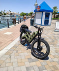 Badass 4000W Dual Motor Electric Mountain Bike For Adults Rip Through Trails At 46 MPH Powered By A Massive 52V 70Ah Battery Yielding Over 200 Mile Range On 26X4 Inch Fat Tires With Full Suspension parked beside a waterfront.