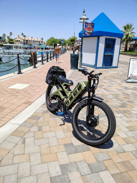 Badass 4000W Dual Motor Electric Mountain Bike For Adults Rip Through Trails At 46 MPH Powered By A Massive 52V 70Ah Battery Yielding Over 200 Mile Range On 26X4 Inch Fat Tires With Full Suspension parked beside a waterfront.