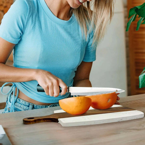 Marble-and-wood cutting board with handle; person slices orange halves on kitchen counter