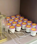 Two Pack 24 Slot Flat Countertop Coffee Pod Holder And Drawer Organizer For Office And Kitchen, Clear Acrylic K Cup Tray Compatible With KCup Pods For Home with filled coffee pods on a shelf next to glassware.