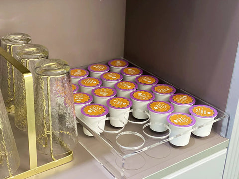 Two Pack 24 Slot Flat Countertop Coffee Pod Holder And Drawer Organizer For Office And Kitchen, Clear Acrylic K Cup Tray Compatible With KCup Pods For Home with filled coffee pods on a shelf next to glassware.