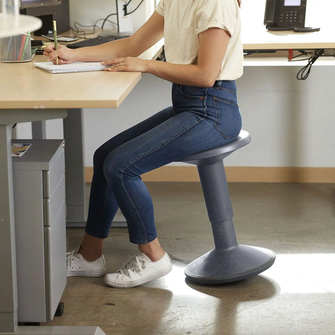 Person seated on SitWell Wobble Stool With Adjustable Height For Active Seating In Classrooms And Libraries, gray finish, office setting.