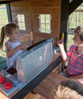 Children playing with kitchen in Cedar Wood Swing Set With Wave Slide And Elevated Clubhouse Play Kitchen And Picnic Area Belt And Web Swings Rock Wall Balconies Playhouse Step Ladder And Outdoor Family Playset For Backyard Fun And Imaginative Outdoor Playtime