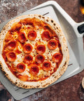 14 Inch Perforated Pizza Peel With Long Handle Aluminum Lightweight Pizza Paddle holding a pepperoni pizza on a stone oven