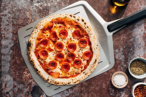 14 Inch Perforated Pizza Peel With Long Handle Aluminum Lightweight Pizza Paddle holding a pepperoni pizza on a stone oven