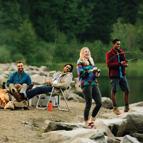 Group of friends fishing by a lakeside with rods, sitting and standing on rocks, surrounded by forest.