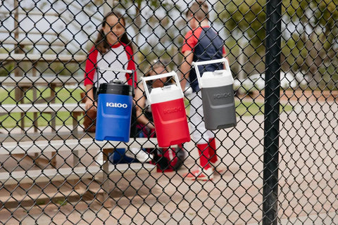 Half-Gallon Pro Blue Beverage Cooler with Secure Flip Cap hangs on a fence, next to red and gray coolers.