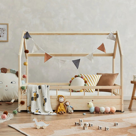 Wooden house-shaped children's bed with decorative pillows, blanket, and toys, including a plush caterpillar and monkey doll.
