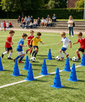 Children playing soccer on a field with blue training cones and footballs, spectators in background watching the game.