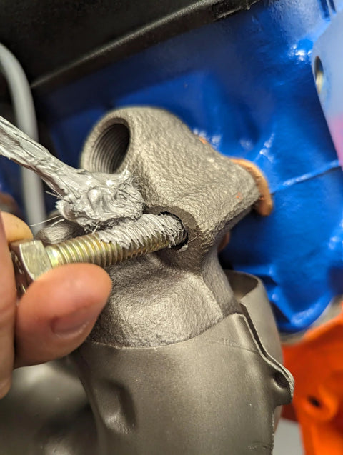 Copper anti-seize lubricant; close-up of threaded bolt entering a gray cast automotive housing against a blue background.