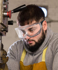 Worker wearing oversized safety goggles that fit over glasses, using a metalworking machine, showcasing anti-fog and splash protection.