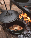 Heavy Duty Dutch Oven Lid Lifter With Coil Handle lifting lid over fire with baked goods inside, outdoor cooking scene
