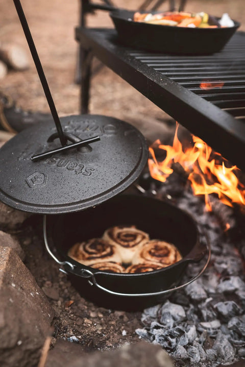 Heavy Duty Dutch Oven Lid Lifter With Coil Handle lifting lid over fire with baked goods inside, outdoor cooking scene