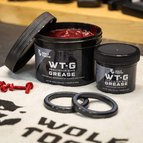Wolf Tooth WT-G Precision Bike Grease containers, 2 oz and 8 oz, on a workshop table with components visible.