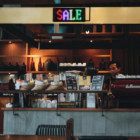 Coffee shop counter with barista, sale sign above, assorted products, and coffee machine under ambient lighting.