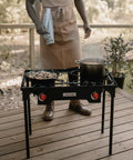 Propane Double Burner Camp Stove With High Heat Output Adjustable PSI Regulator 4ft Hose Removable Legs Great For Outdoor Cooking Tailgating And Catering Use on a wooden deck with a person cooking.