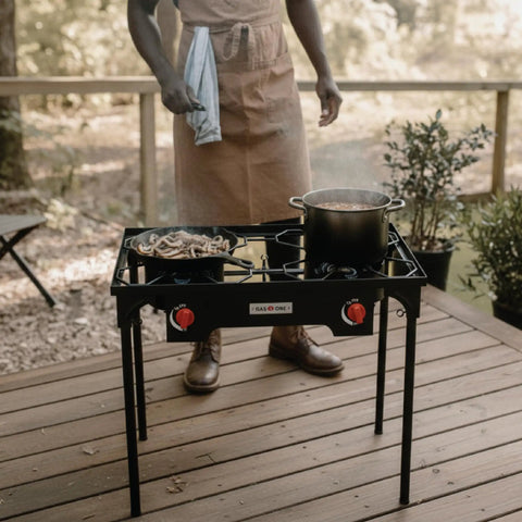 Propane Double Burner Camp Stove With High Heat Output Adjustable PSI Regulator 4ft Hose Removable Legs Great For Outdoor Cooking Tailgating And Catering Use on a wooden deck with a person cooking.