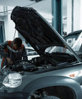 Front view of auto technician inspecting engine bay with raised hood on a silver SUV in a workshop