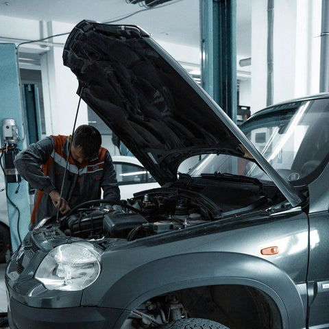 Front view of auto technician inspecting engine bay with raised hood on a silver SUV in a workshop