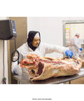 Butcher in white coat and gloves cuts a large beef carcass on a stainless steel table in a meat processing facility.
