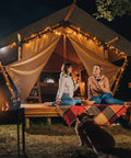 Two people sitting outside an illuminated tent at night, with a fire pit and guitar nearby, enjoying a relaxed outdoor setting.