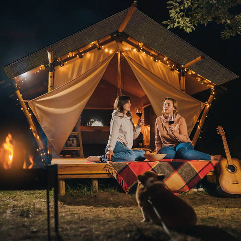 Two people sitting outside an illuminated tent at night, with a fire pit and guitar nearby, enjoying a relaxed outdoor setting.
