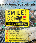 Smile You're On Camera Signs Two Pack Outdoor Video Surveillance Warning Signs 10x7 Rust Free Aluminum UV Printed Weatherproof Security Sign on a chain-link fence.