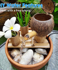 DIY water feature setup with bamboo and coconut components, flowing water into a stone-filled basin, plumeria flower displayed.
