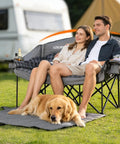Couple sitting on Beast Mode Double Camping Chair with cup holders, grey color, dog resting on carry bag mat, outdoor setting.