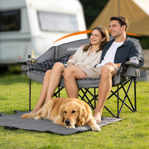 Couple sitting on Beast Mode Double Camping Chair with cup holders, grey color, dog resting on carry bag mat, outdoor setting.
