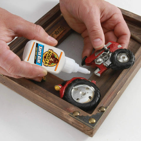 Hands using Fast-Drying Thick-Bond Duo Pack Clear Super Glue For Metal Wood Ceramic Rubber And More, Quick Set In 10-45 Seconds, Anti-Clog Cap, Two 15 Gram Bottles For Easy Home Repairs to repair a toy tractor on a wooden surface