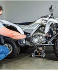 Man using Triple Lift 3000 Pound Heavy Duty Garage Jack For Cars Trucks Motorcycles And Transmissions With 4.75 To 17 Inch Lift Range Wide Wheelbase For Stability Includes Safety Bar And Locking Mechanism to lift an ATV in a garage.