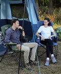 Two adults sitting outside a tent, one in an oversized folding high back lawn chair for adults with cup holder, carrying strap, hard armrests, 400lb capacity, blue, ideal for camping, fishing, hunting, sports, and backyard; blue chair visible