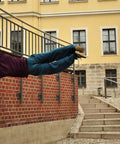 Person performing parkour move by hanging sideways from railing outside a building with brick wall and staircase.
