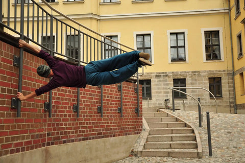 Person performing parkour move by hanging sideways from railing outside a building with brick wall and staircase.