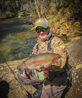Person holding a large trout by a river, wearing camouflage clothing and a hat, with fishing rod nearby.