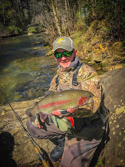 Person holding a large trout by a river, wearing camouflage clothing and a hat, with fishing rod nearby.