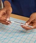 Hands holding a utility knife with Number 11 Fine Point Utility Blades Pack Of 100 Ready For Precise Cutting And Trimming, on a grid mat.