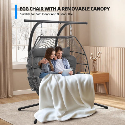 Couple sitting in an oversized double hanging swing chair with removable canopy and cushion indoors, enjoying a tablet.