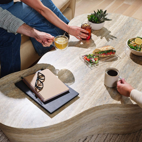 Marble coffee table with notebooks, glasses, drinks, salad, and sandwich with people reaching for beverages.
