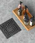 6 Inch Matte Black Shower Drain Square Floor Drain Kit With Flange Stainless Steel Pro Grade Construction Removable Grid Cover Hair Strainer High Flow CUPC Certified Fits 2 Inch Central Outlet next to a soap bottle tray on a tiled floor