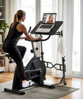 Woman using exercise bike with standing desk on wheels for treadmills with adjustable height 29.5 to 45.3 inches, laptop on surface.