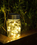 Hanging Mason Jar Solar Lights 6 Pack With 30 LED Lanterns glowing on a wooden ledge in a garden, foliage background.