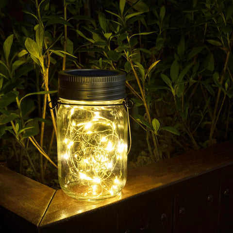 Hanging Mason Jar Solar Lights 6 Pack With 30 LED Lanterns glowing on a wooden ledge in a garden, foliage background.