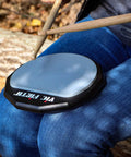 Six Inch Single Sided Practice Pad With Realistic Drumhead Feel And Low Volume For Quiet Jamming On The Go Natural Rubber On A Durable Wood Base To Train Wrists And Keep Your Chops Tight, shown on lap with drumsticks